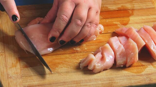 A knife in a woman's hand cuts a chicken breast on a wooden cutting board. Close-up.