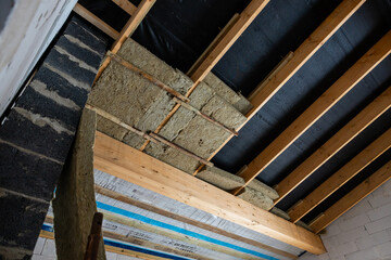 Ceiling area of a building under construction, featuring wooden beams, partially installed insulation, and a concrete block wall on the left.