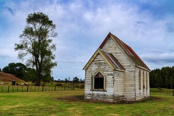 Obraz premium Abandoned church on a rural property. Horses graze nearby. A weathered, white wooden structure, with a faded red metal roof. Ruawai, Dargaville, Northland, NZ