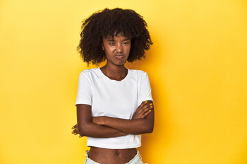 Teen girl in classic white T-shirt, yellow studio backdrop frowning face in displeasure, keeps arms...