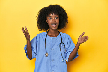 Young African-American nurse in studio with yellow background receiving a pleasant surprise,...