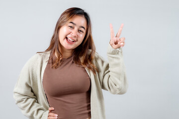 A cheerful young woman in a cozy sweater makes a peace sign, radiating positivity and a playful spirit against a neutral background.