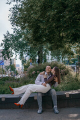 Happy couple in love enjoying a playful moment in the city park. A young man sitting on a bench holding his girlfriend in his arms as they smile at each other. Urban nature, romance, and joy.