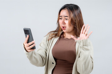 Fototapeta premium A young woman expresses frustration while looking at her smartphone, showcasing a strong emotional reaction in a neutral background.