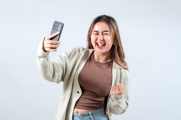 Fototapeta premium A young woman celebrates joyfully while holding her smartphone, expressing excitement and triumph in a neutral background.