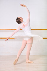 Young female ballerina with brunette hair elegantly posing in ballet studio. Dressed in white leotard, embracing classic form against backdrop of wooden barre and light walls © TRAVELARIUM