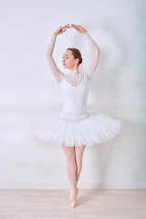 Naklejka premium Young female ballerina in elegant white tutu stands gracefully on pointe in bright minimalist studio. Soft lighting highlights serene expression and delicate posture against light-toned background