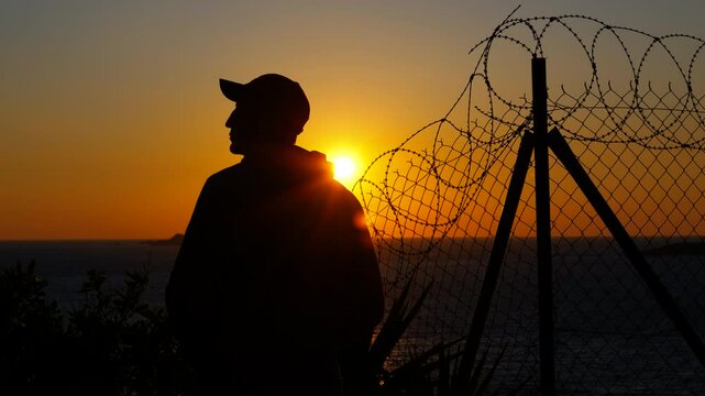 Refugee observing sunset behind barbed wire fence. Man silhouetted in baseball cap standing near barbed wire fence, gazing at golden sunset over expansive ocean horizon, embodying themes of freedom