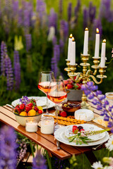 Two glasses of rose wine, flowers in a vase, silverware, fruits. Sunset, golden hour, atmospheric candlelight. Romantic table decor for a loving couple on blooming meadow with purple lupines