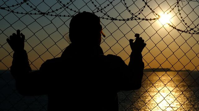 Refugee at sunset near the border with barbed wire. Silhouette of a refugee woman at sunset near the sea coast looks around in fear