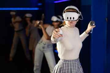 Young woman fully immersed in game using VR headset and controllers, standing in futuristic dark blue gaming room, actively gesturing to manipulate objects in virtual world