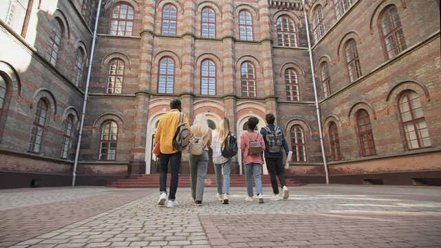 Camera view from behind of group of students walking towards college campus main entrance. Holding notebooks and wearing backpacks. Big old building with many windows and ornaments on walls.