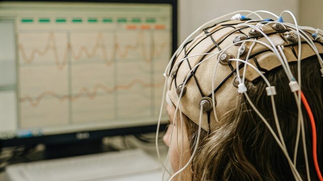 A detailed view of an EEG cap on a human head with electrodes attached to the scalp and data streaming on a nearby monitor visually representing the bridge between neural signals