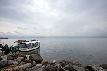 A serene scene at Lake Sapanca, Sakarya, Turkey, features a white boat docked beside a rocky shore. The water is calm, reflecting the overcast sky filled with grey clouds.