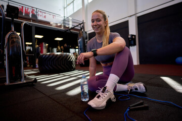 Happy athlete woman taking a break after exercising with jump rope at fitness club, sitting on floor with bottle of water and checking smartwatch