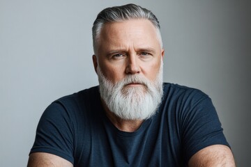 Fototapeta premium Older man with a gray beard and short hair poses against a neutral background in casual attire