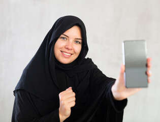 smiling Muslim woman in traditional black hijab shows blank smartphone screen close-up. Dark touch...