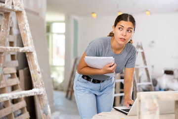Fototapeta premium Young woman designer checking documents and using her laptop during repair works indoors.