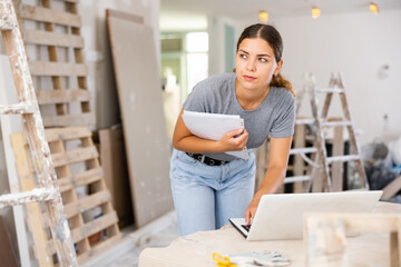 Female caucsaian architect holding documents, using laptop during repair works in house.