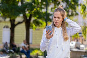 Fototapeta premium Caucasian young woman using smartphone typing browsing, loses becoming surprised sudden lottery results, bad news, fortune loss, fail. Tourist girl standing in urban street outdoors. Town lifestyles.