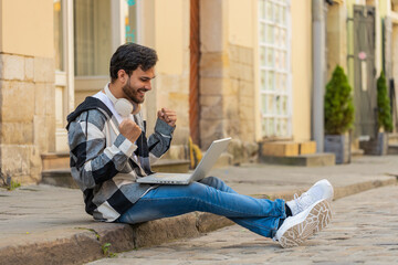 Handsome Hispanic young man using laptop computer celebrating win good message news, lottery jackpot victory, giveaway online outdoors. Happy Indian guy tourist sitting on city street. Town lifestyles