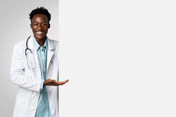 Smiling black male doctor pointing at big blank advertisement board with open palm, professional...