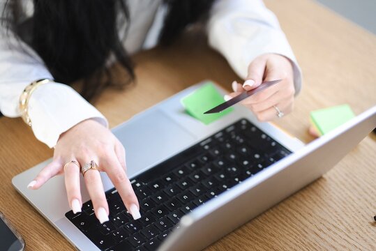 A close-up of a woman in a white shirt holding a credit card while using a laptop, indicating an online shopping or banking transaction in a workspace setting.