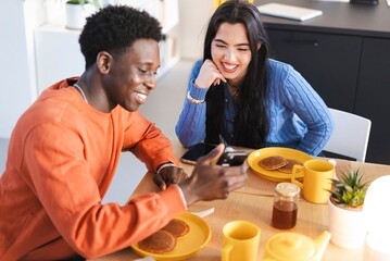 A man in an orange sweater and a woman in a blue sweater smile while sharing a breakfast of pancakes and coffee at a cozy dining table, engaging in a cheerful conversation.