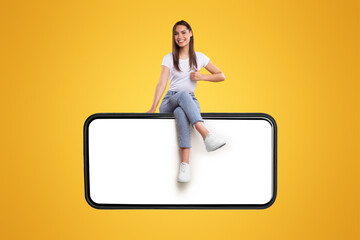 Cheerful young woman sitting on big huge horizontal new cellphone with white empty screen and showing thumb up gesture, mockup, casual lady enjoying newest entertaining mobile app, orange wall