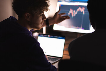 A young man in a purple hoodie is seen in profile, gesturing towards a computer screen displaying financial charts, engaged in a discussion with a colleague in a dimly lit office.