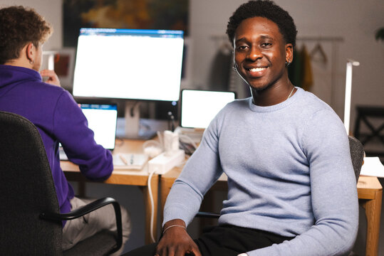 A young man in a light blue sweater sits at a modern desk, smiling at the camera, with financial charts on monitors and a colleague in the background focused on work.