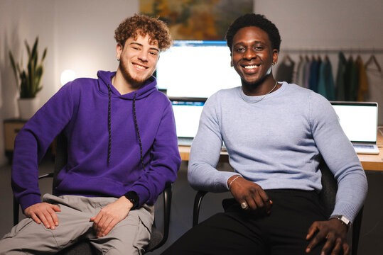 Two young men, one wearing a purple hoodie and the other in a blue sweater, sit together at a desk in a modern office setting with financial charts on screens in the background.