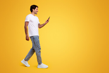 Communication, Technology And Lifestyle Concept. Full body length of smiling young guy using cell phone, walking isolated on orange studio background, profile side view panoramic banner, free space © Prostock-studio