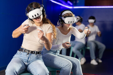 Girls in a VR helmet with headphones and joysticks are actively playing virtual game against guys....