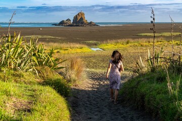 Woman in floral dress walks along a sandy path at a beach. Coastal scenery. Whatipu, Auckland, New...