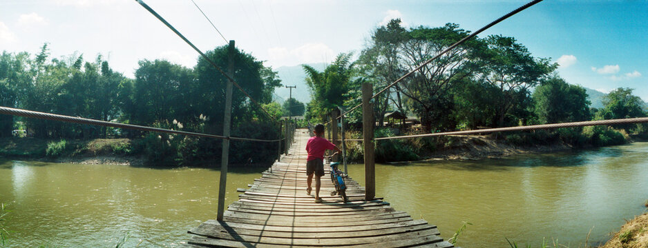 Panoramic view of child walking with his bike across an old wooden bridge over the river, Chiang Mai Province, Thailand.