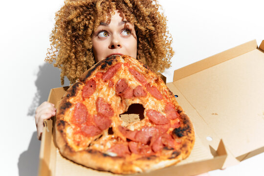 A cheerful young woman with curly hair holding a large pepperoni pizza, expressing excitement and joy, showcasing the concept of indulgent eating and sharing food