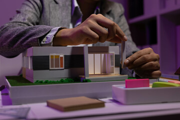 A woman is sitting at a desk with two model houses in front of her. She is holding a pencil and she...