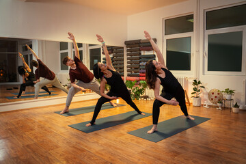 Group workout on yoga mat, two women and young man in sync performing yoga stretches, night time in...