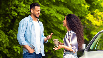 Joyful multiracial millennial couple having coffee break at countryside while car trip, standing by auto, drinking tea and having conversation, discussing details of their journey, free space