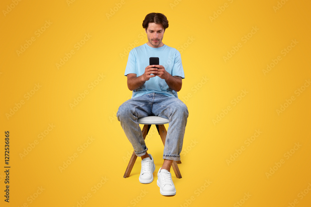 Poster full body length vertical portrait of focused millennial man using phone sitting in chair isolated o