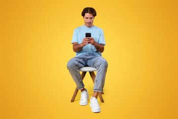 Full Body Length Vertical Portrait Of Focused Millennial Man Using Phone Sitting In Chair Isolated Over Orange Studio Background. Serious Guy Using New Mobile Application On Smartphone, Checking Email