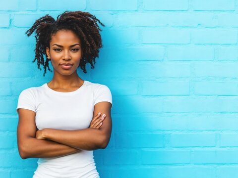 Mulher negra de bra&ccedil;os cruzados em fundo azul de tijolo, com express&atilde;o s&eacute;ria e confiante