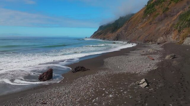 Waves crashing surf black sand rugged coastline sunny morning blue sky mist fog layer USAL Beach Campground Lost Coast Trail California aerial drone PNW boulders rocks mountainside upwards motion