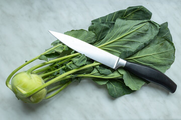 A fresh kohlrabi with green leaves lies on a marble surface, accompanied by a sharp kitchen knife...