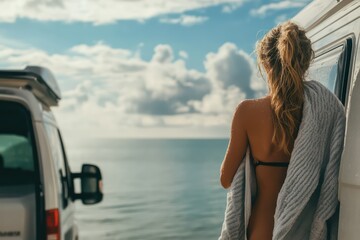 Fototapeta premium A woman with wet hair in a towel, standing next to her camper van with the sea and sky in the distance, enjoying her time by the water after a swim.