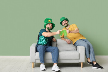 Happy young men in leprechaun hats with green beards holding glasses of beer and sitting on sofa...