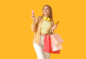 Beautiful young happy woman with headphones, credit cards and shopping bags on yellow background