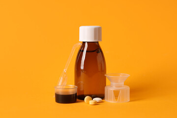Bottle of cough syrup with measuring cups and pills on orange background