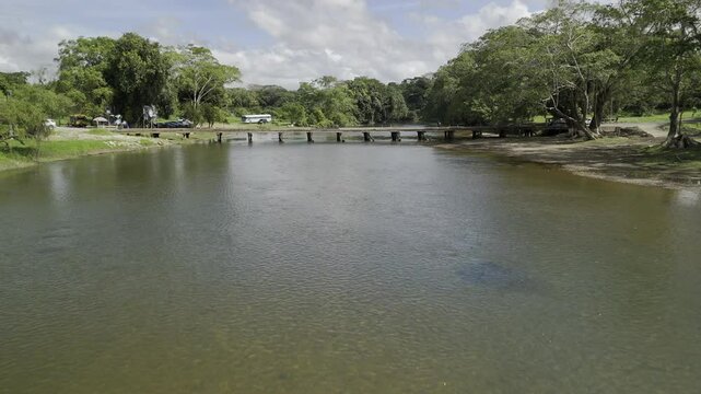 Drone flies north low over Macal River lined with lush trees, crossing over small wooden bridge in San Ignacio, Belize